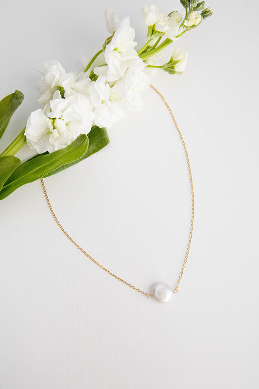A freshwater coin pearl hung on a 14k gold filled chain necklace lays on a white background with a white flower running across the top of the image.