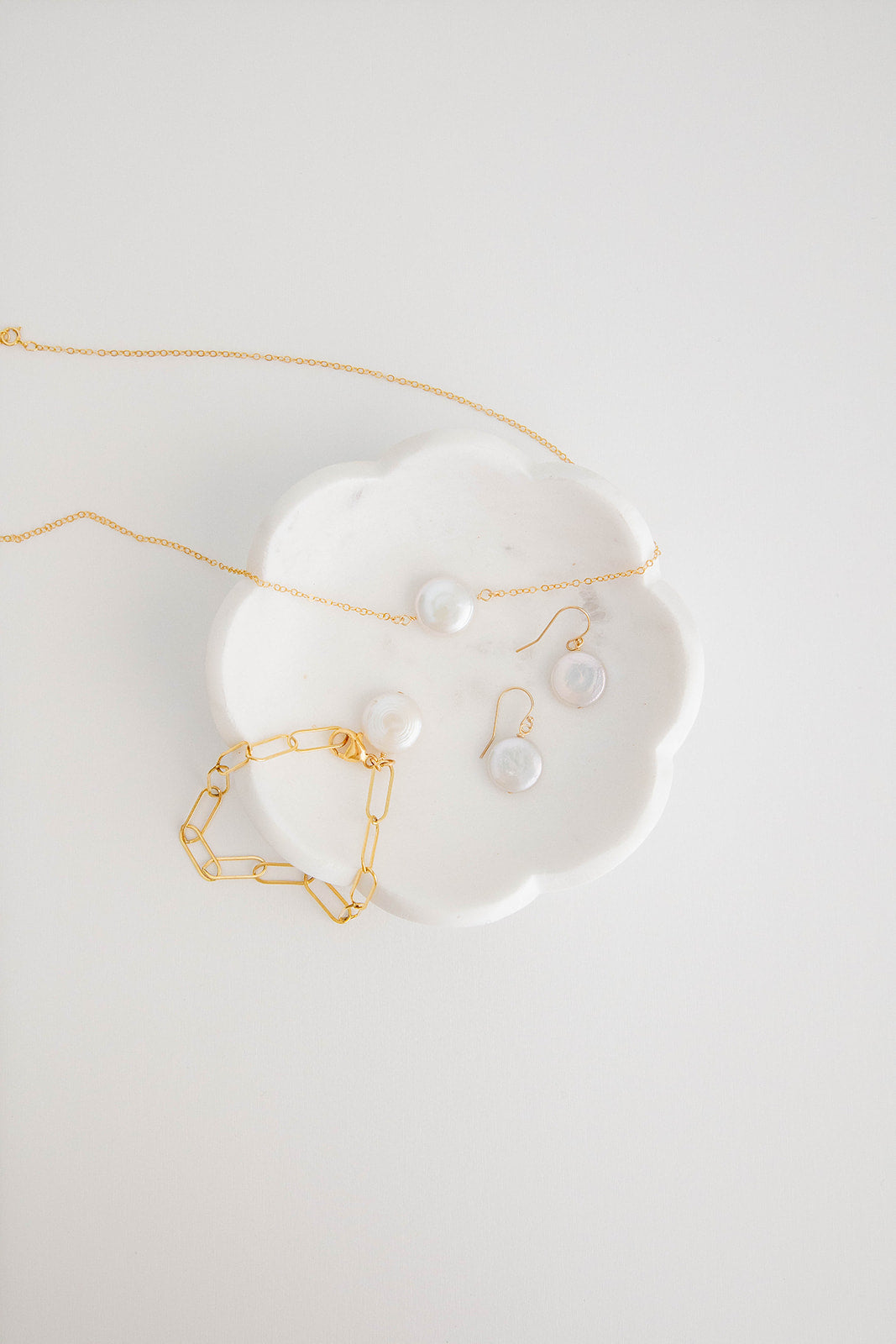 A pair of earrings with freshwater pearl coins hung on gold filled shepherd’s hooks lay on a white marble dish on a white flatlay background with matching necklace and bracelets.