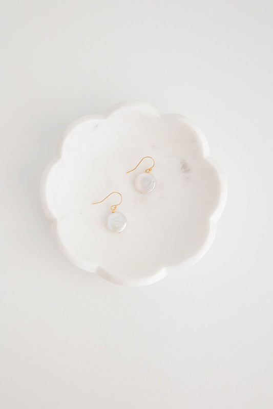 A pair of earrings with freshwater pearl coins hung on gold filled shepherd’s hooks lay on a white marble dish on a white flatlay background.