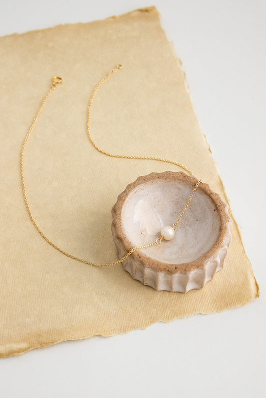A round freshwater pearl hangs on a 14k gold filled chain necklace on a brown textured paper flatlay.