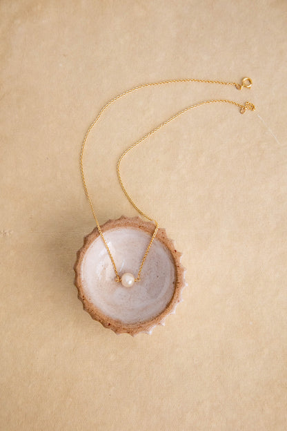 A round freshwater pearl hangs on a 14k gold filled chain necklace sits inside a brown ceramic bowl on a brown textured paper flatlay.