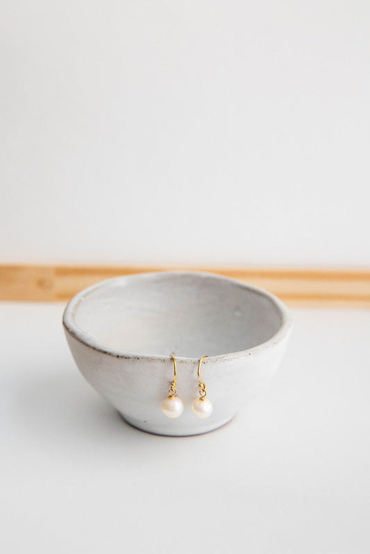 A pair of mini pearl earrings hung on shepherd’s hooks hang from a small white ceramic bowl.