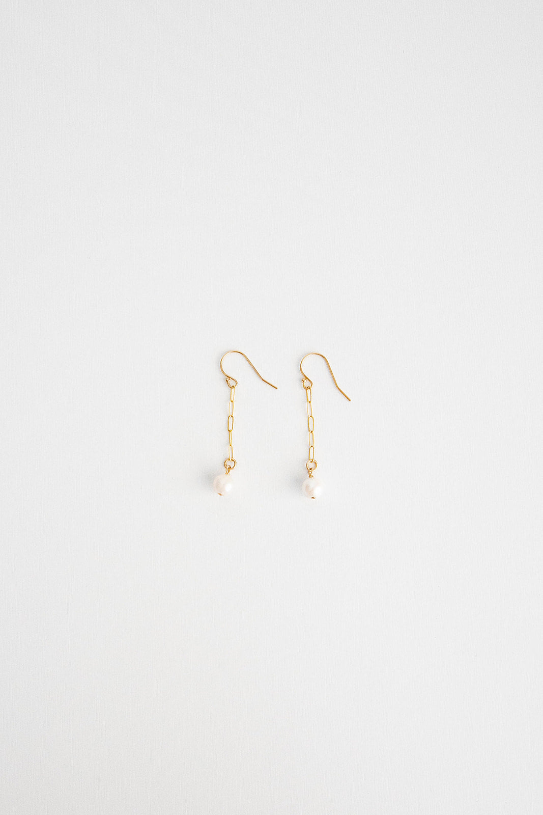 A pair of mini pearl earrings hung on shepherd’s hooks lay on a white background flatlay.