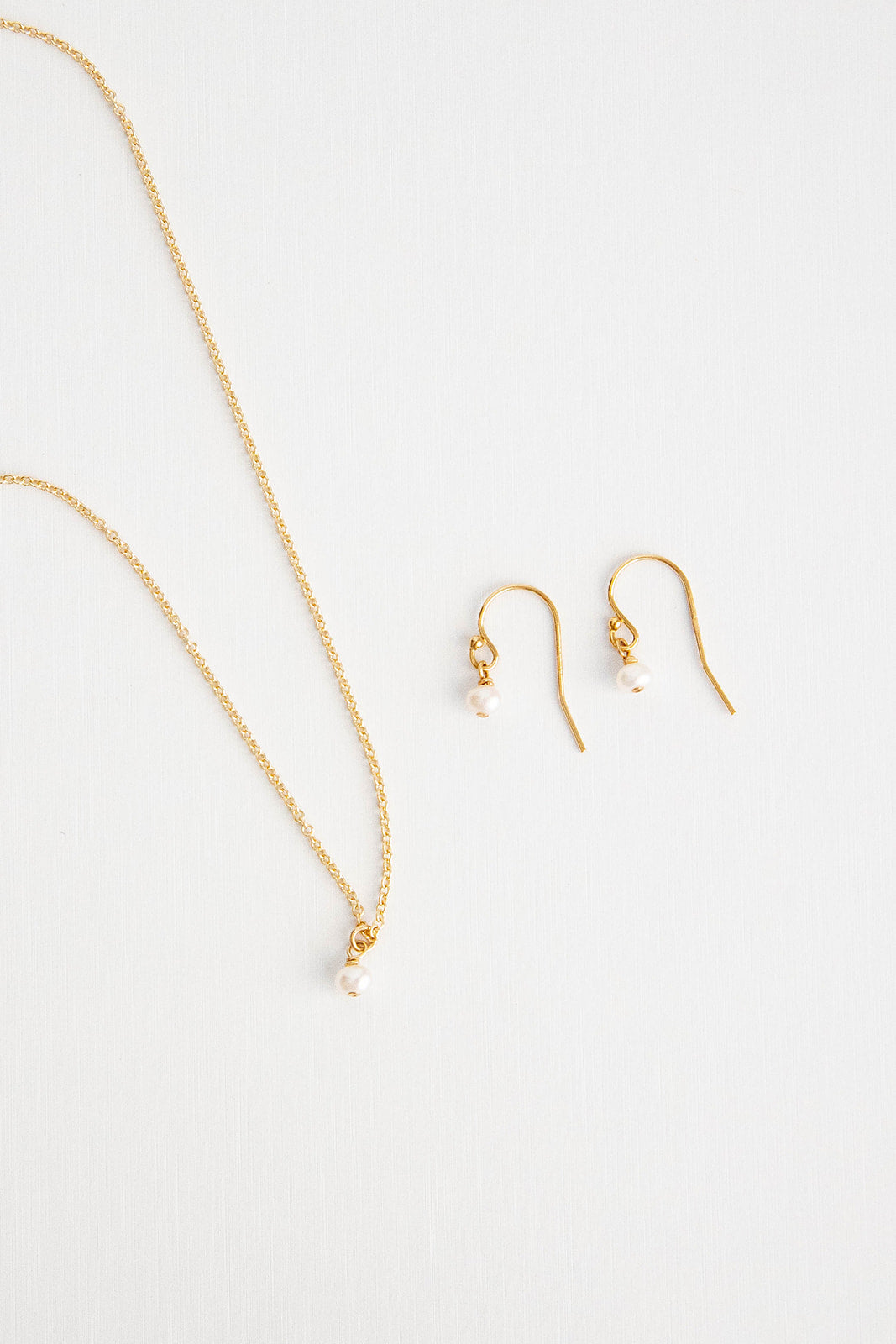 A pair of mini pearl earrings hung on shepherd’s hooks lay on a white background flatlay with matching necklace.