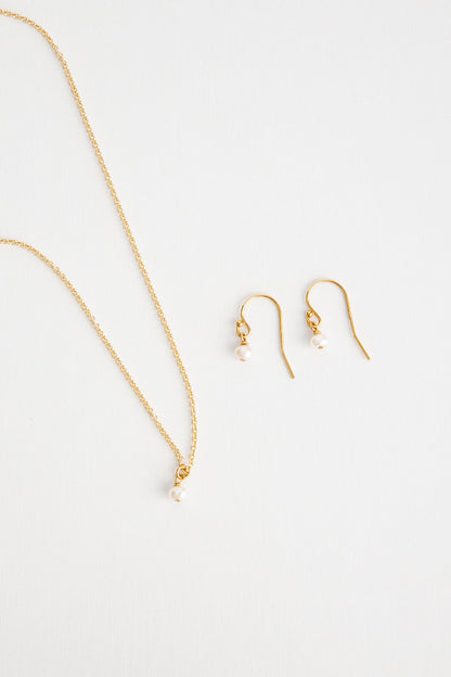A pair of mini pearl earrings hung on shepherd’s hooks lay on a white background flatlay with matching necklace.