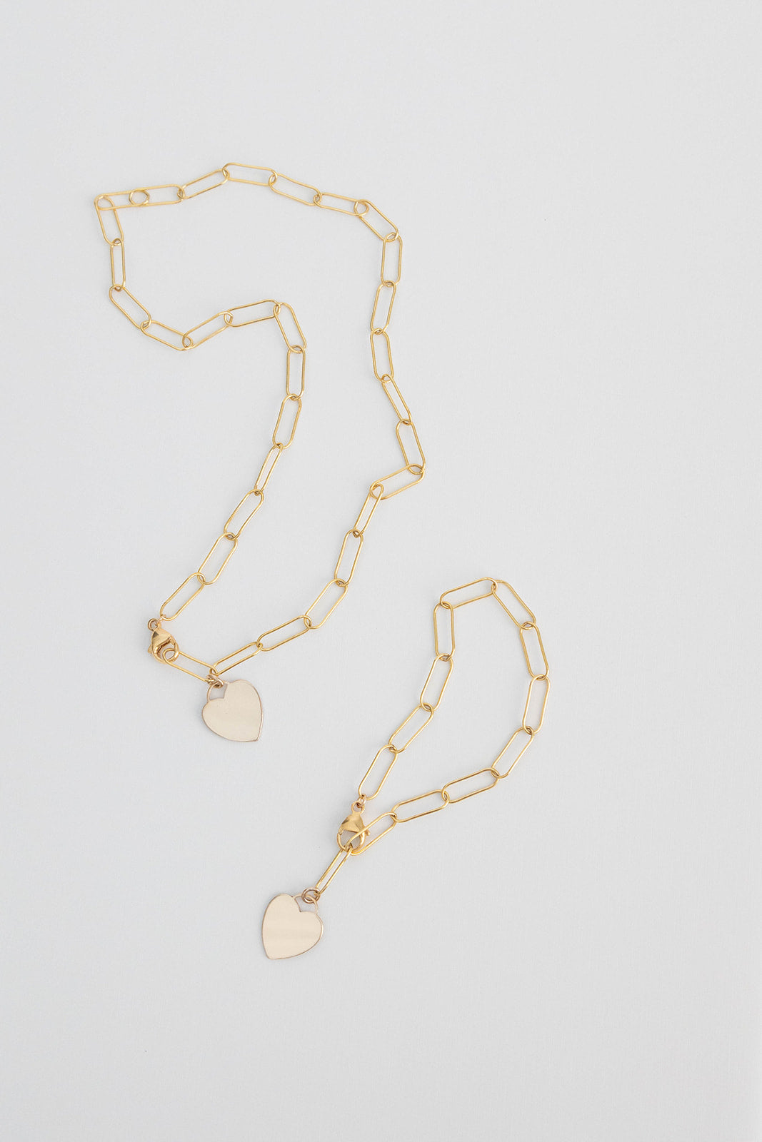 A 7 inch 14k gold filled paper clip chain bracelet with heart tag lays on a white background with a matching necklace.