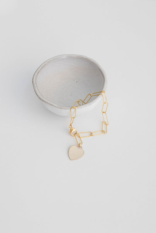 7 inch 14k gold filled paper clip chain bracelet with heart tag lays on a small white ceramic bowl with a white background.