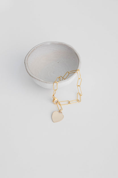 A 14k gold filled paper clip chain bracelet hangs from a small white ceramic bowl on a white background.