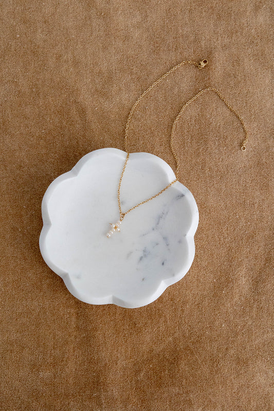 A 14k gold filled cable link chain necklace with freshwater pearls connected in the shape of a cross drape over a white marble dish on a brown textured flatlay background.