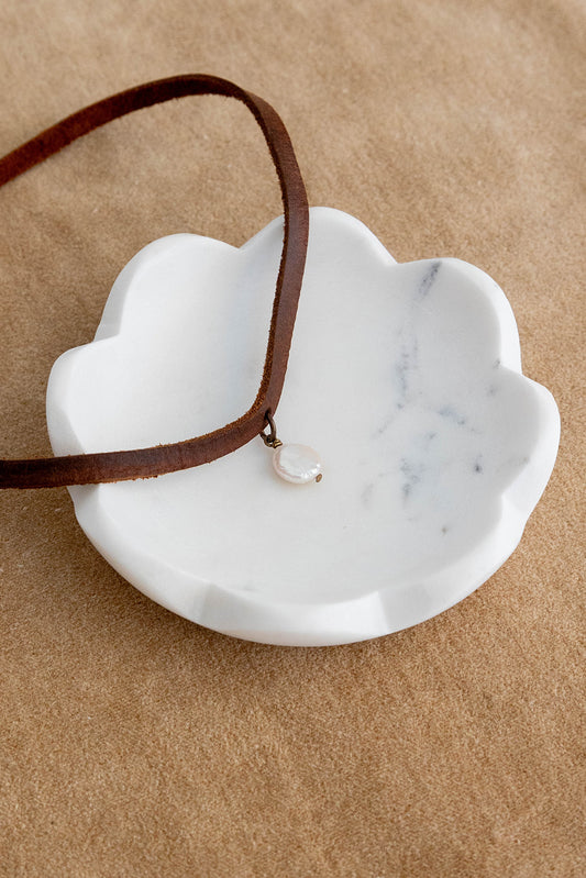A leather strap choker with a freshwater pearl coin lays on a white marble ceramic dish on top of a brown textured background.