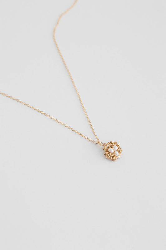 A 14k gold filled wire necklace wrapped to mimic a bird nest with freshwater pearls lays on a white background.