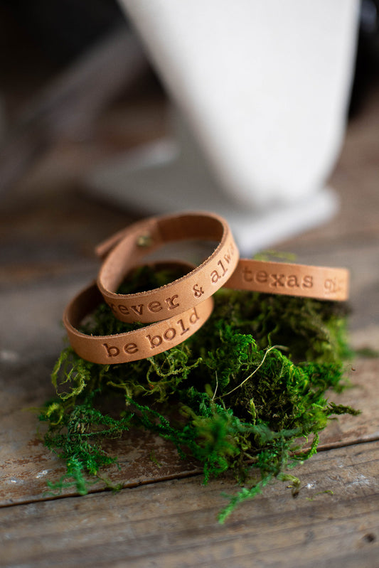 A stack of three hand stamped leather bracelets with knob closure lay on top of green moss for a styled product photo.