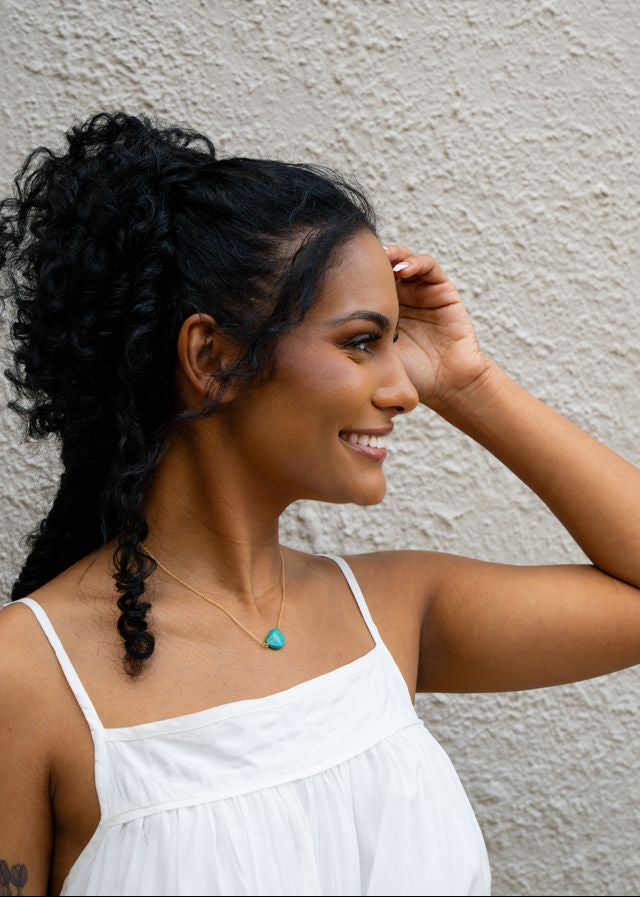 A woman with dark curly hair in a high ponytail wearing a white spaghetti-strap top and a gold necklace with a turquoise pendant, posing against a textured beige stucco wall.