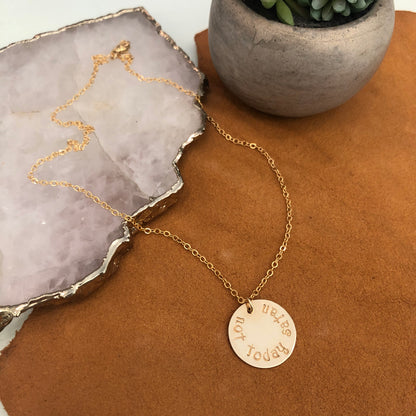 A 14k gold filled chain necklace with a 14k gold filled hand stamped disc reading "not today satan" drapes over a quartz piece onto a brown leather background.