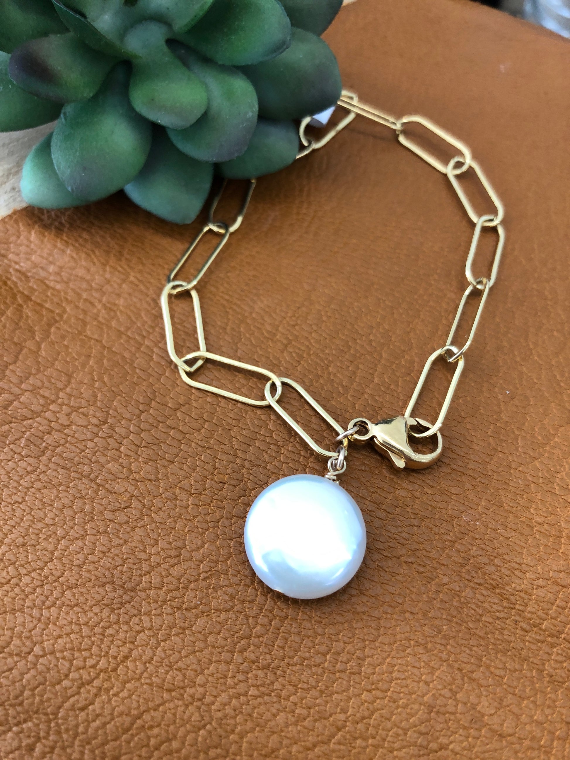 A 14k gold filled paper clip chain bracelet with a “larger than life” freshwater pearl coin lays on a brown leather piece with faux succulent sneaking from the left corner.