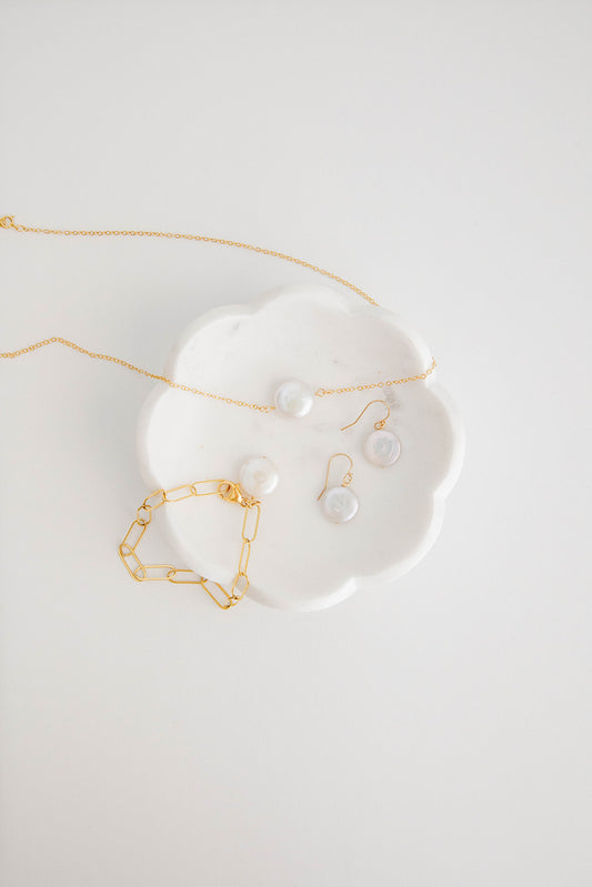 A 14k gold filled paper clip chain necklace with a “larger than life” freshwater pearl coin draped over a white ceramic dish with matching earrings and bracelets.