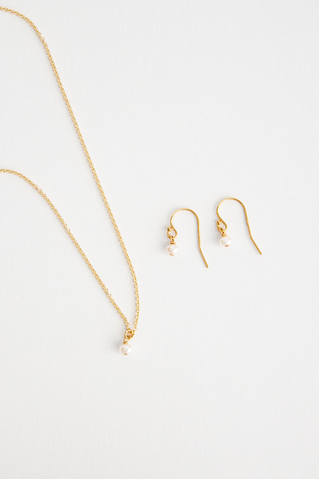 A mini freshwater pearl on a 14k gold filled cable link chain necklace lays next to a pair of matching earrings lays on a white background.