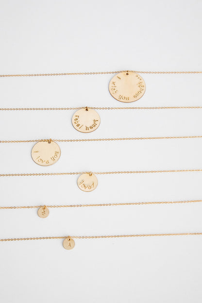 A 25.4 mm 14k gold filled disc hung on a 14k gold cable link chain necklace lays on a white background with more gold necklaces in different sizes.