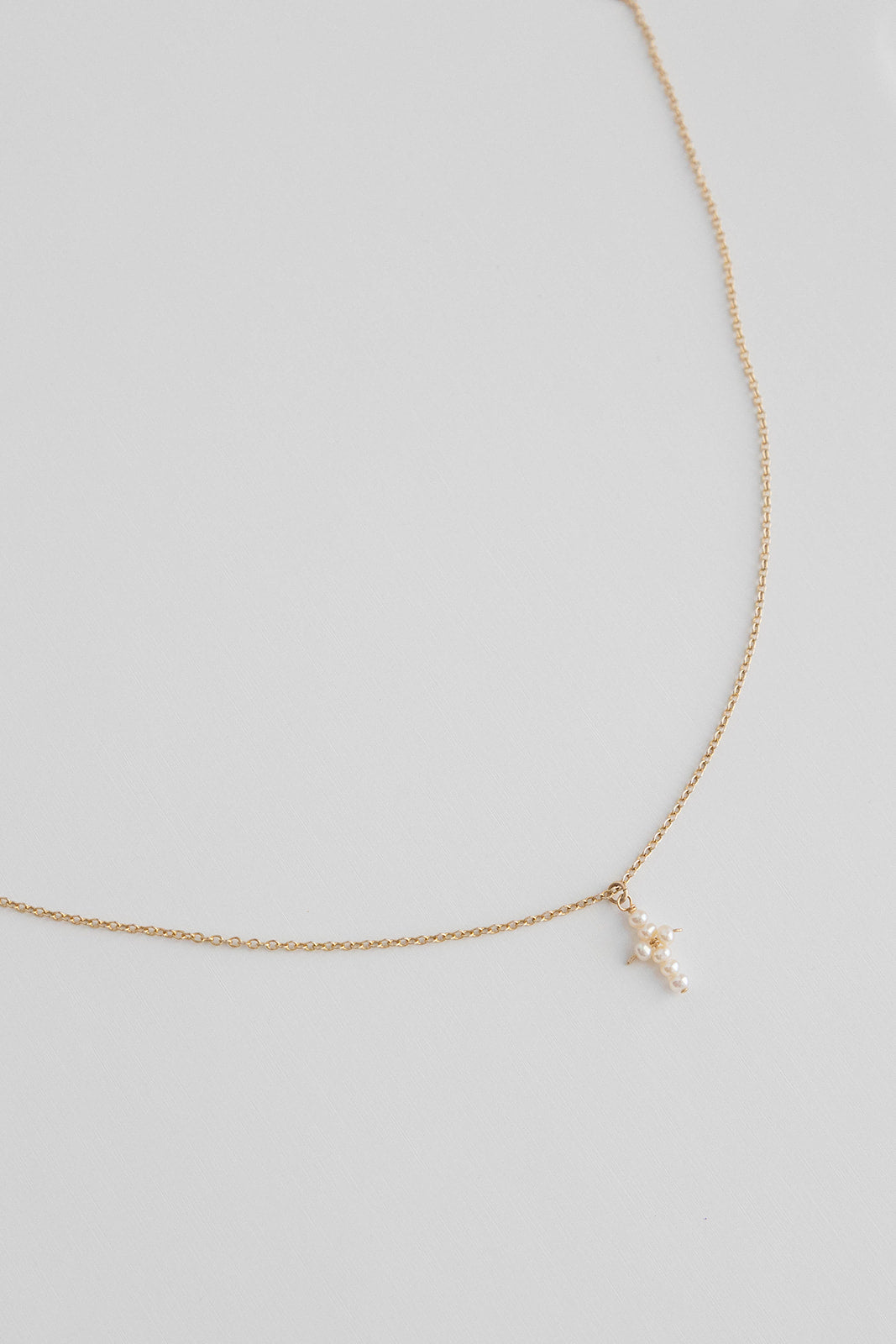 A 14k gold filled cable link chain necklace with freshwater pearls connected in the shape of a cross lays on a white background.