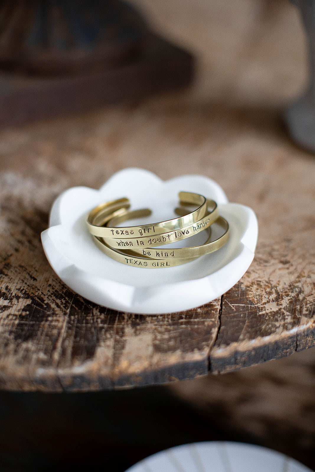A stack of four brass hand stamped cuffs lay on a white marble dish on a wood tabletop.