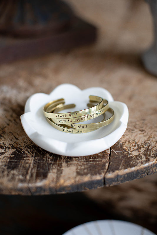 A stack of four brass hand stamped cuffs lay on a white marble dish on a wood tabletop.