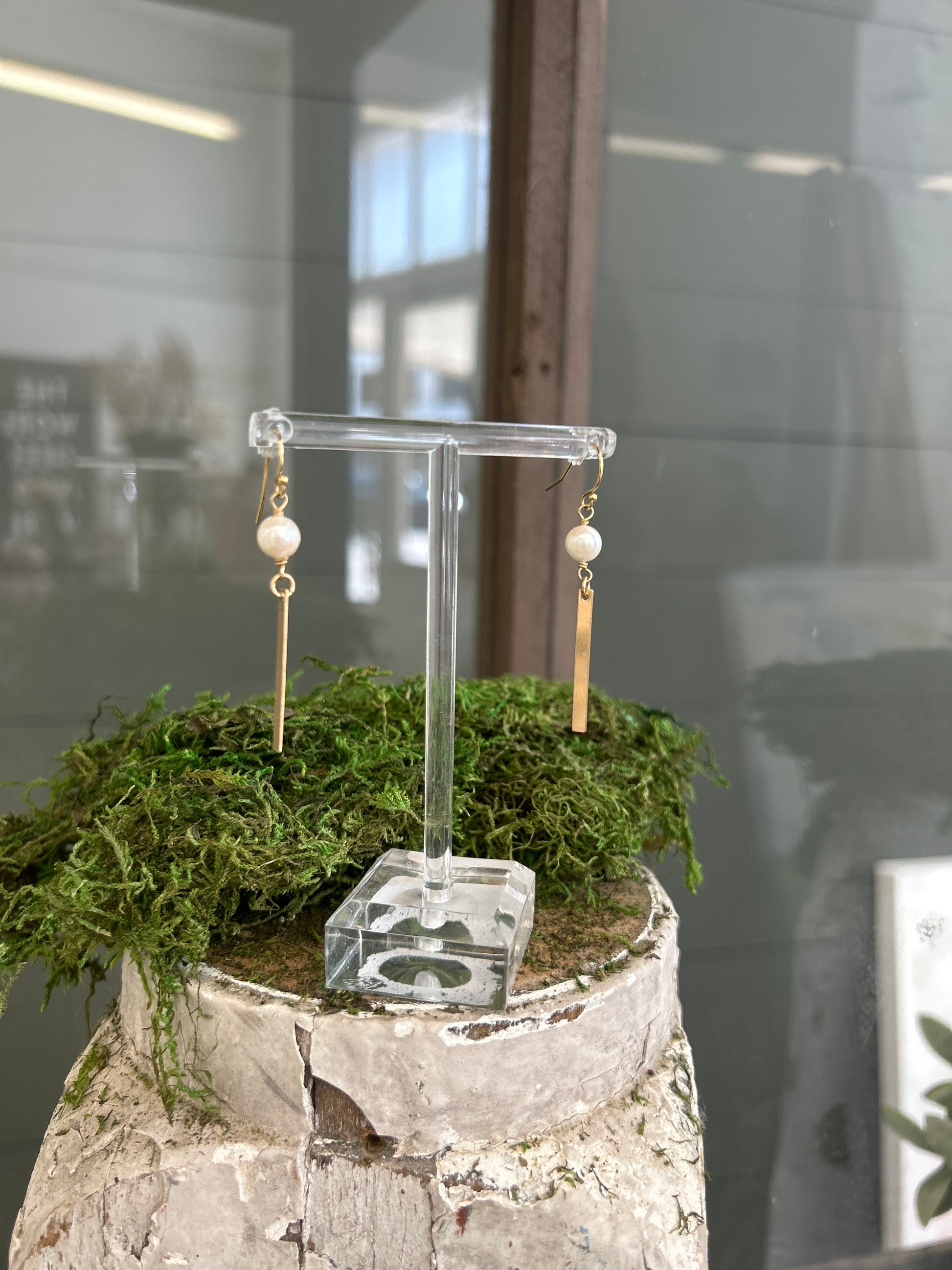 A pair of earrings with freshwater pearls sit above a 14k gold filled bar and hang on shepherd’s hooks on a clear acrylic earring stand.