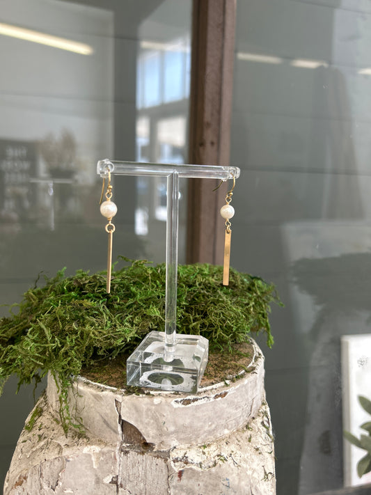 A pair of earrings with freshwater pearls sit above a 14k gold filled bar and hang on shepherd’s hooks on a clear acrylic earring stand.