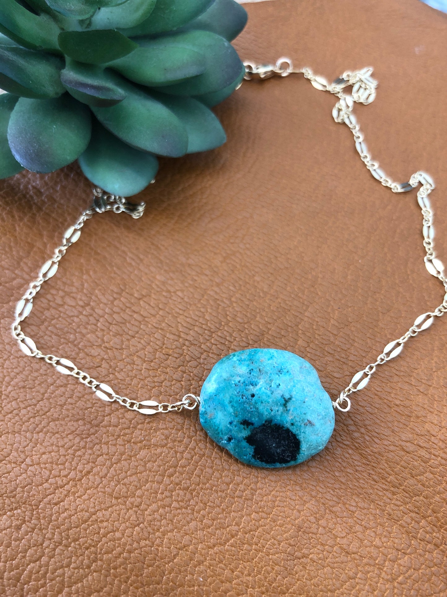 A 14k gold filled 18" lace necklace with a turquoise gemstone lays on a brown leather piece.