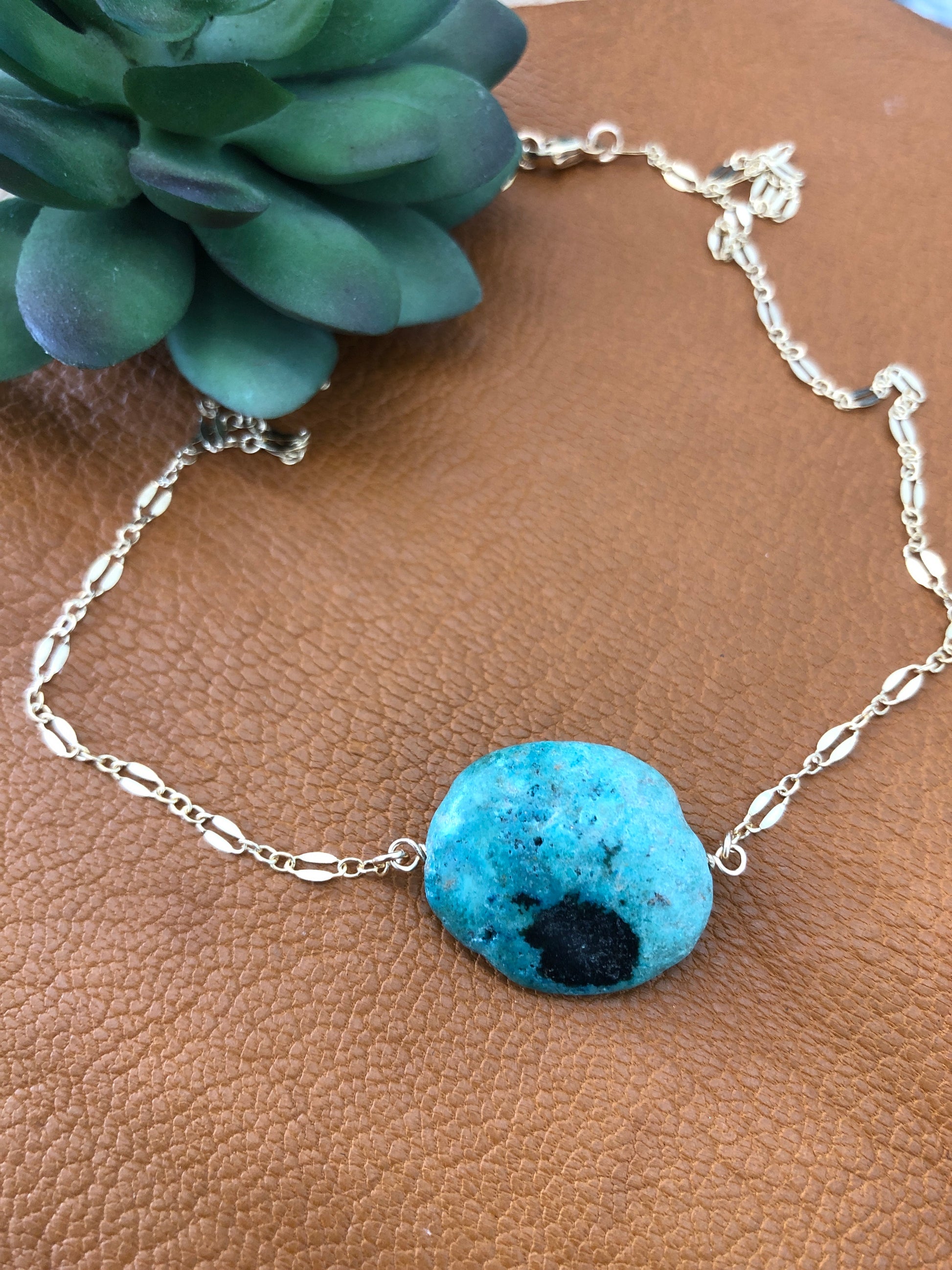 A 14k gold filled 18" lace necklace with a turquoise gemstone lays on a brown leather piece.