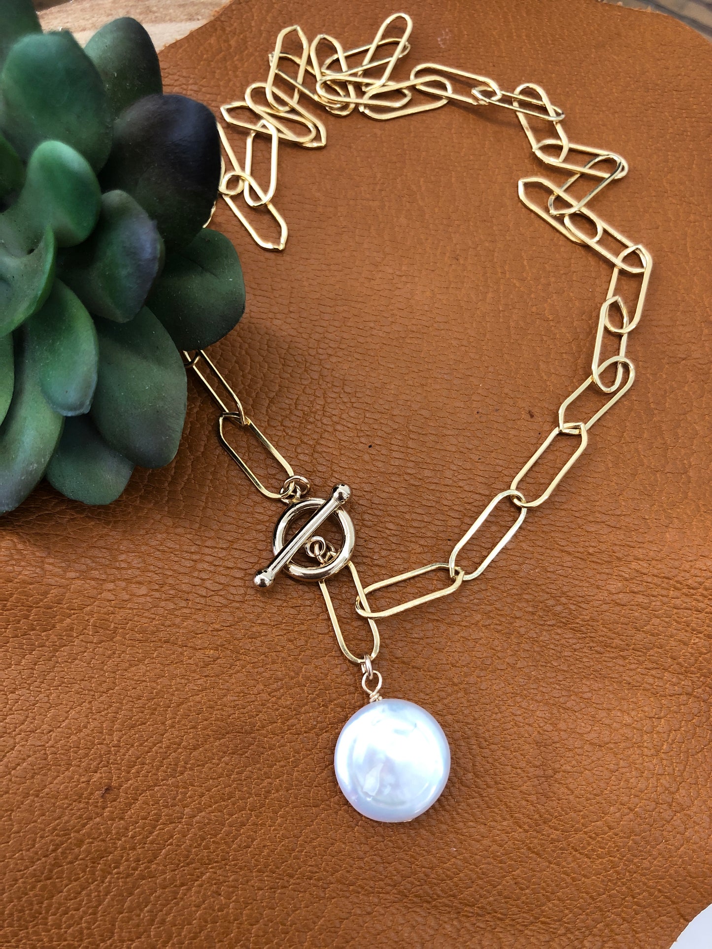 A 14k gold filled large paper clip necklace with fresh water coin pearl and toggle lays on a brown leather piece with a faux succulent sitting on the top left corner of the image.