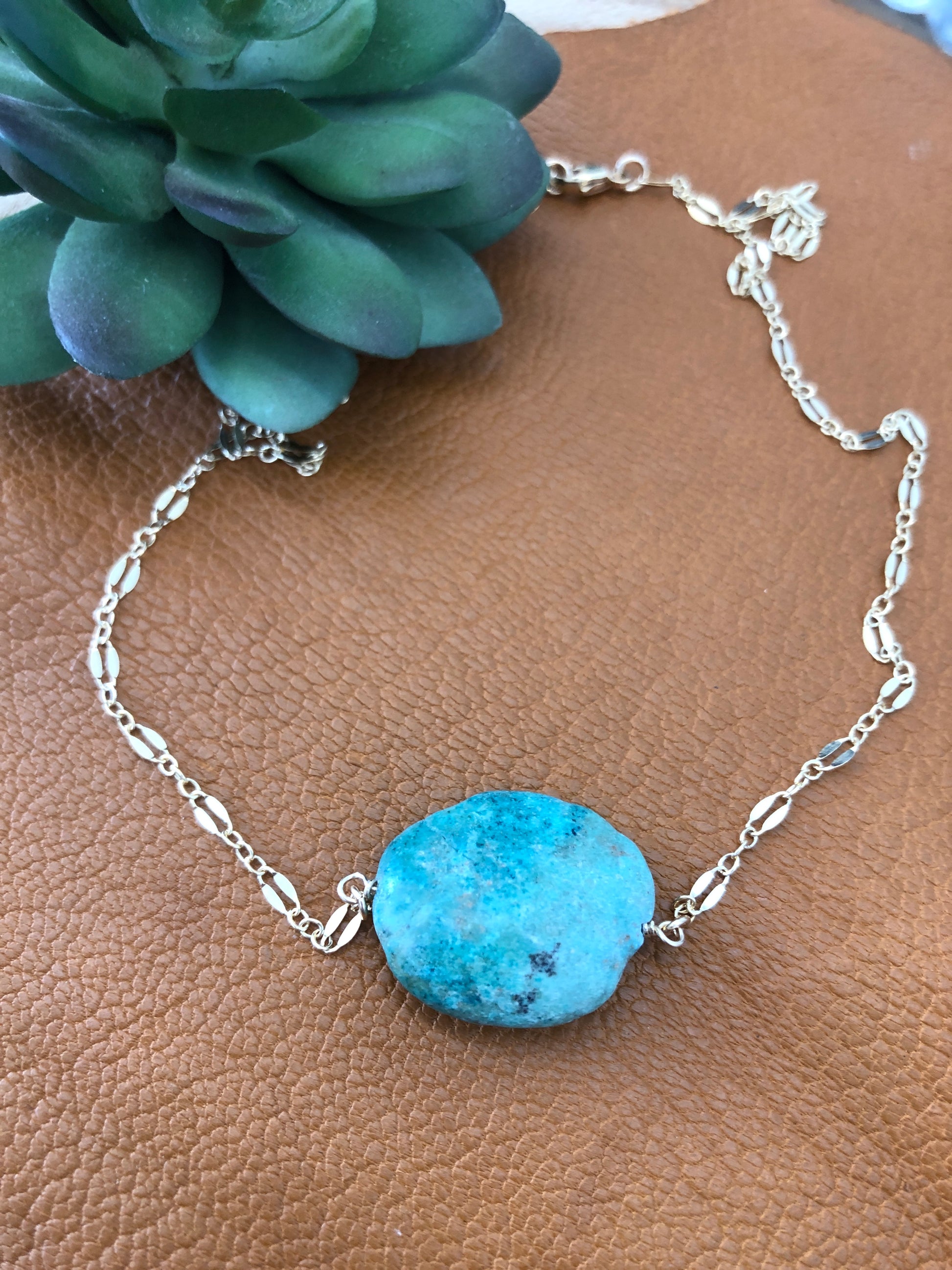 A 14k gold filled 18" lace necklace with a turquoise gemstone lays on a brown leather piece.